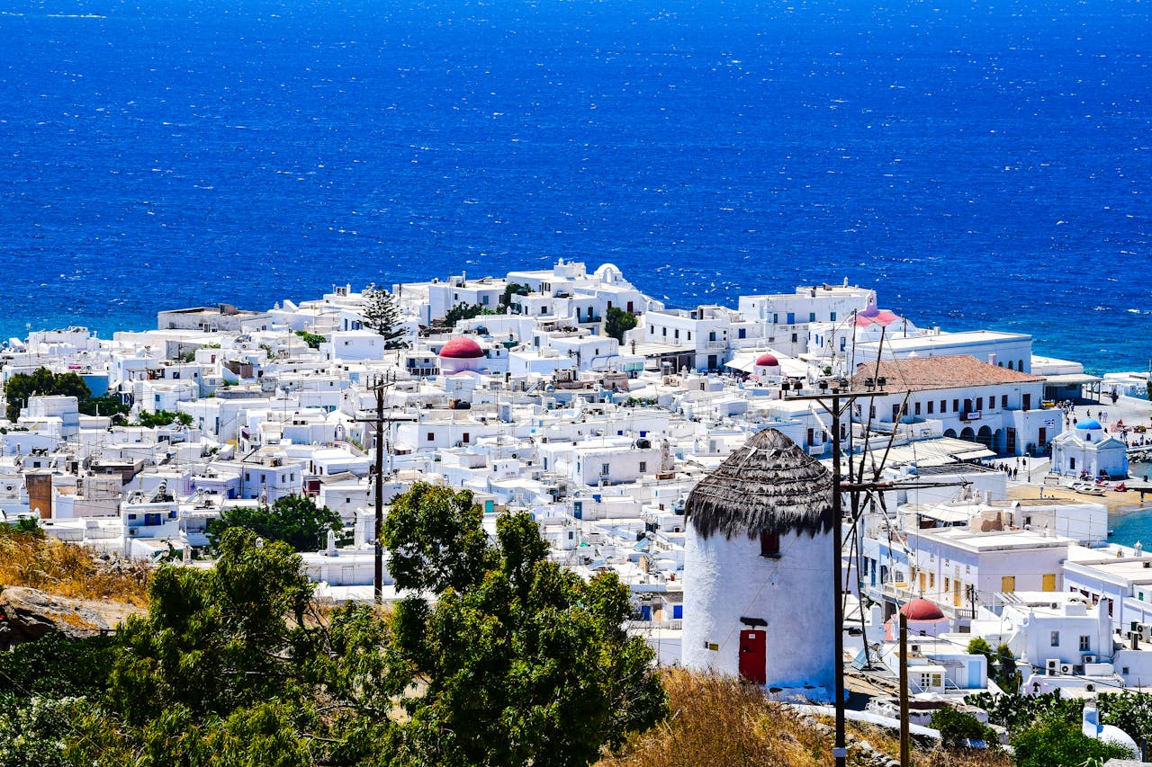 High-angle view of whitewashed Mykonos Town houses and a traditional thatched windmill against the blue Aegean Sea in Greece.