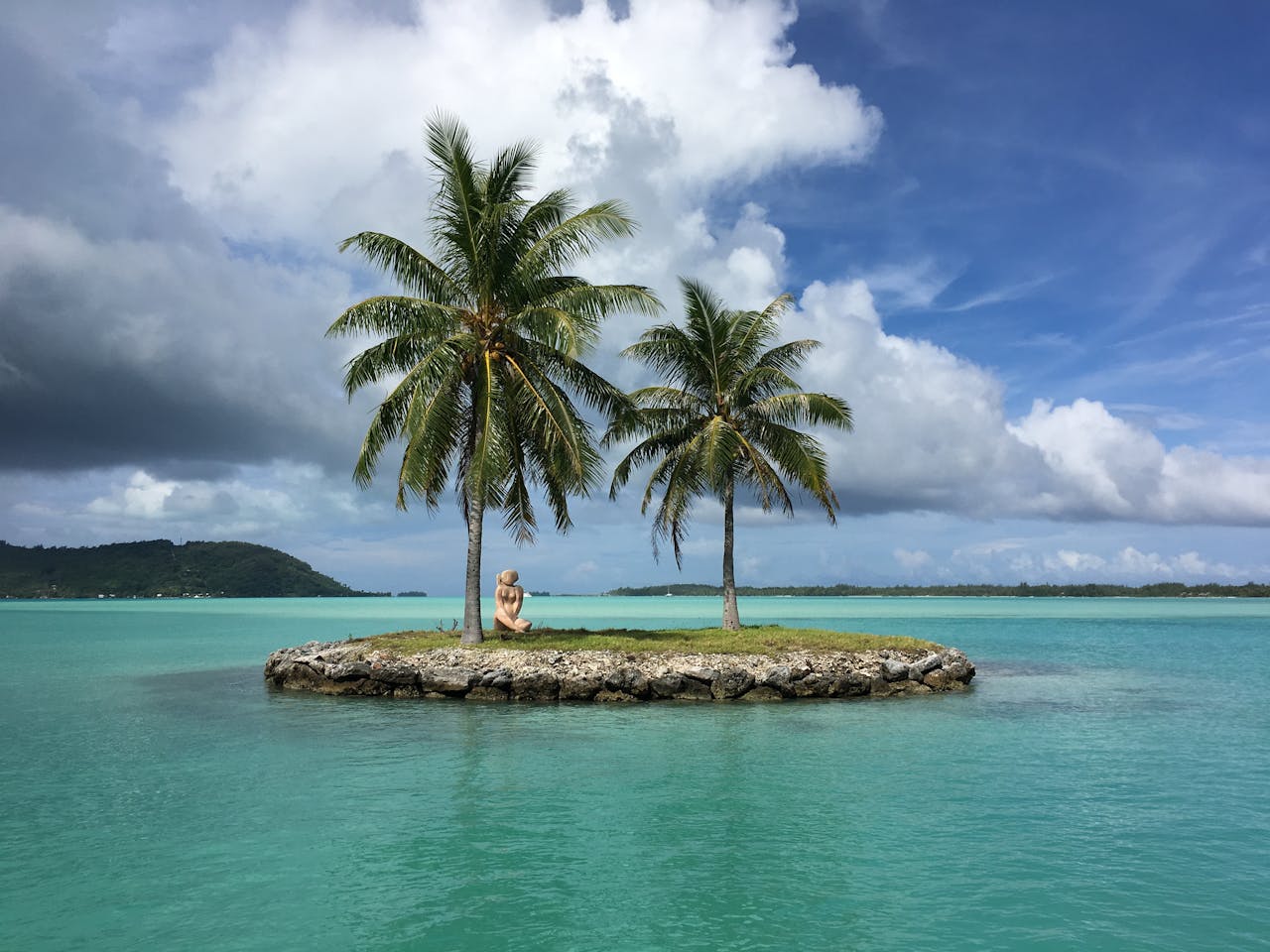 A tiny tropical islet with two palm trees and a Tiki statue in the turquoise lagoon of Bora Bora, French Polynesia.